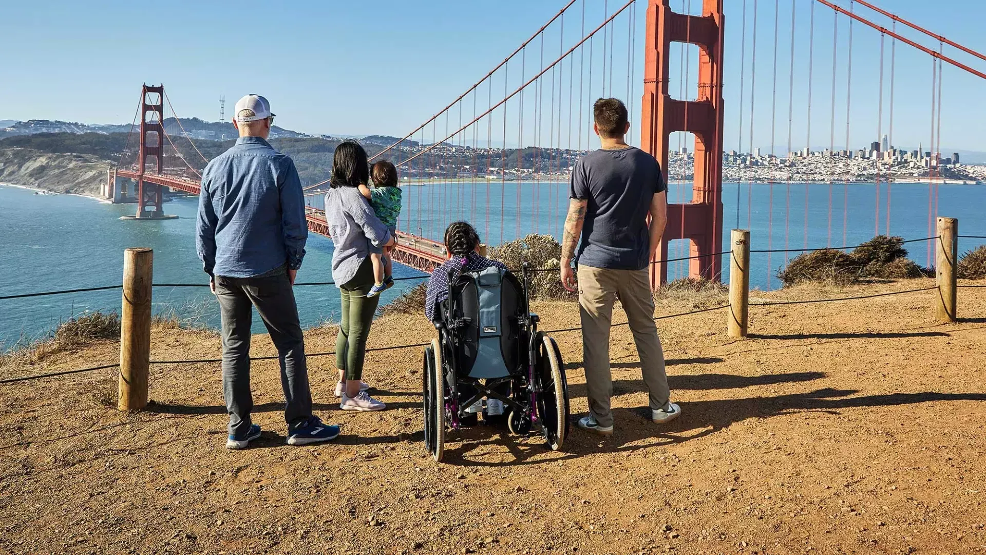 Eine Gruppe von Menschen, darunter eine Person im Rollstuhl, ist von hinten zu sehen, wie sie von den Marin Headlands aus auf die Golden Gate Bridge blicken.